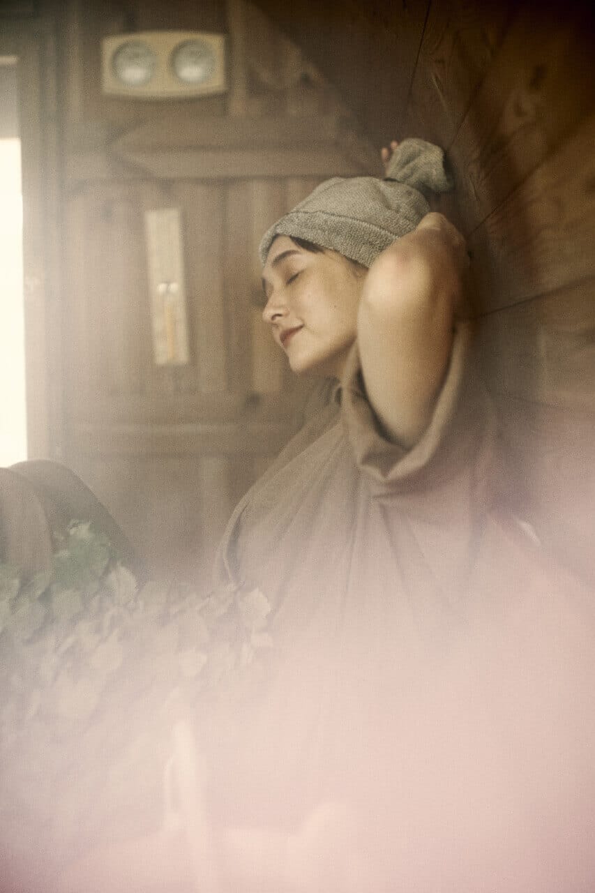Woman relaxing in a wooden sauna, wearing a towel and knit cap with eyes closed.