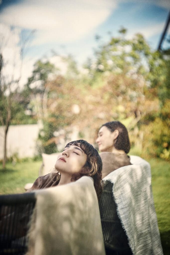 Two women relax in a sunny garden on outdoor lounge chairs, one with eyes closed and a blanket over her shoulders while the other sits nearby reading.