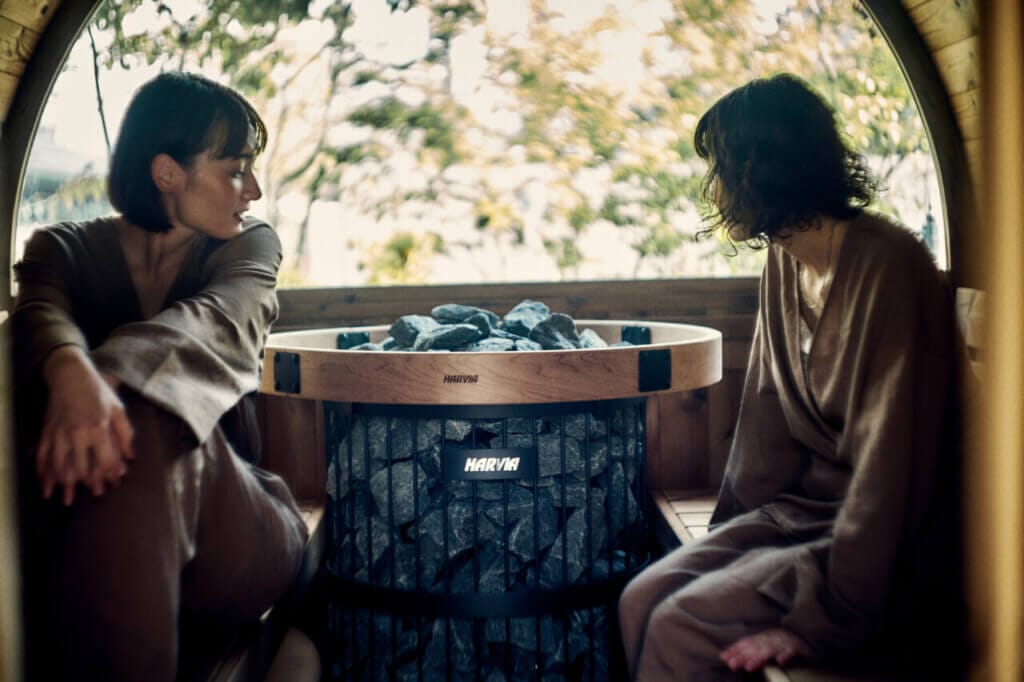 Two women sit in a round wooden barrel sauna with a rock heater labeled Harvia, gazing to the side.