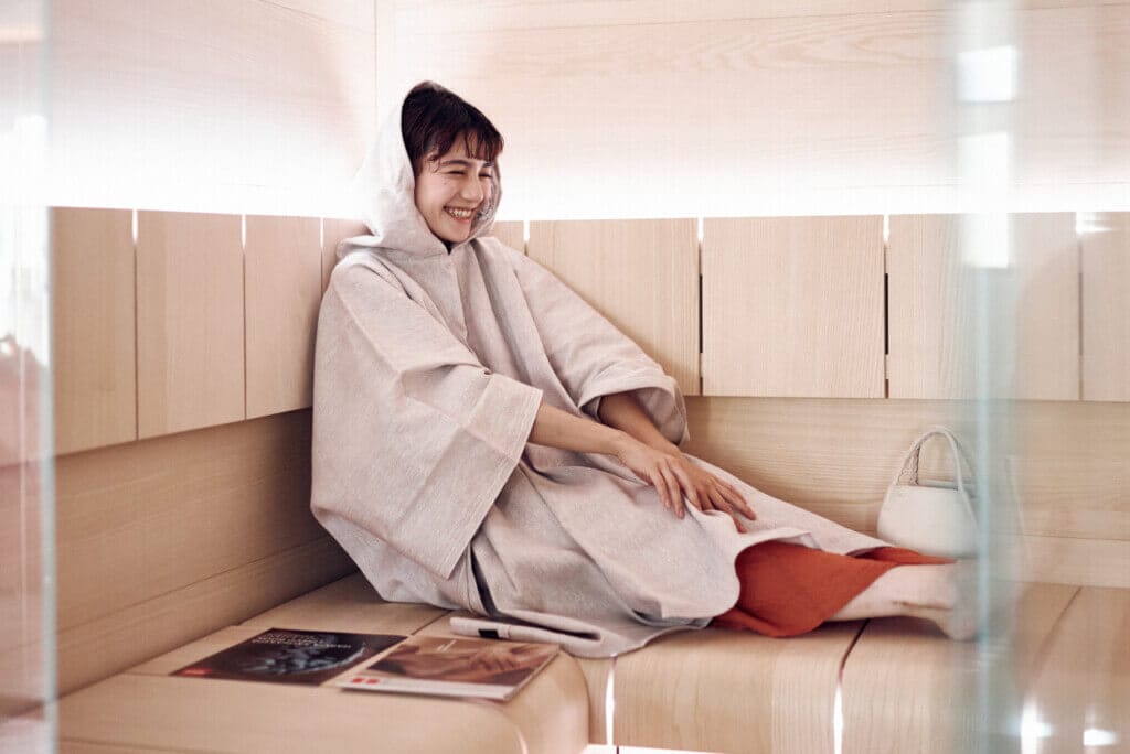 Person in a light robe sits on a wooden bench in a spa sauna, smiling contentedly with a white bag beside them.
